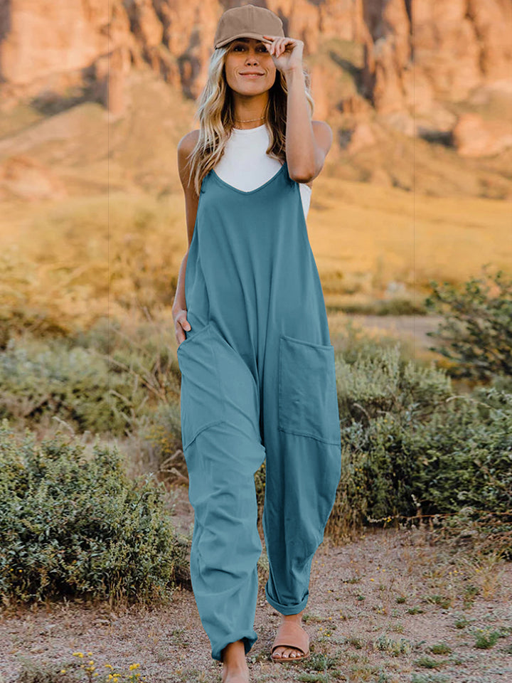 Person wearing a Double Take Full Size V-Neck Sleeveless Jumpsuit with Pockets and sandals, walking outdoors on a dirt trail with bushes in the background.