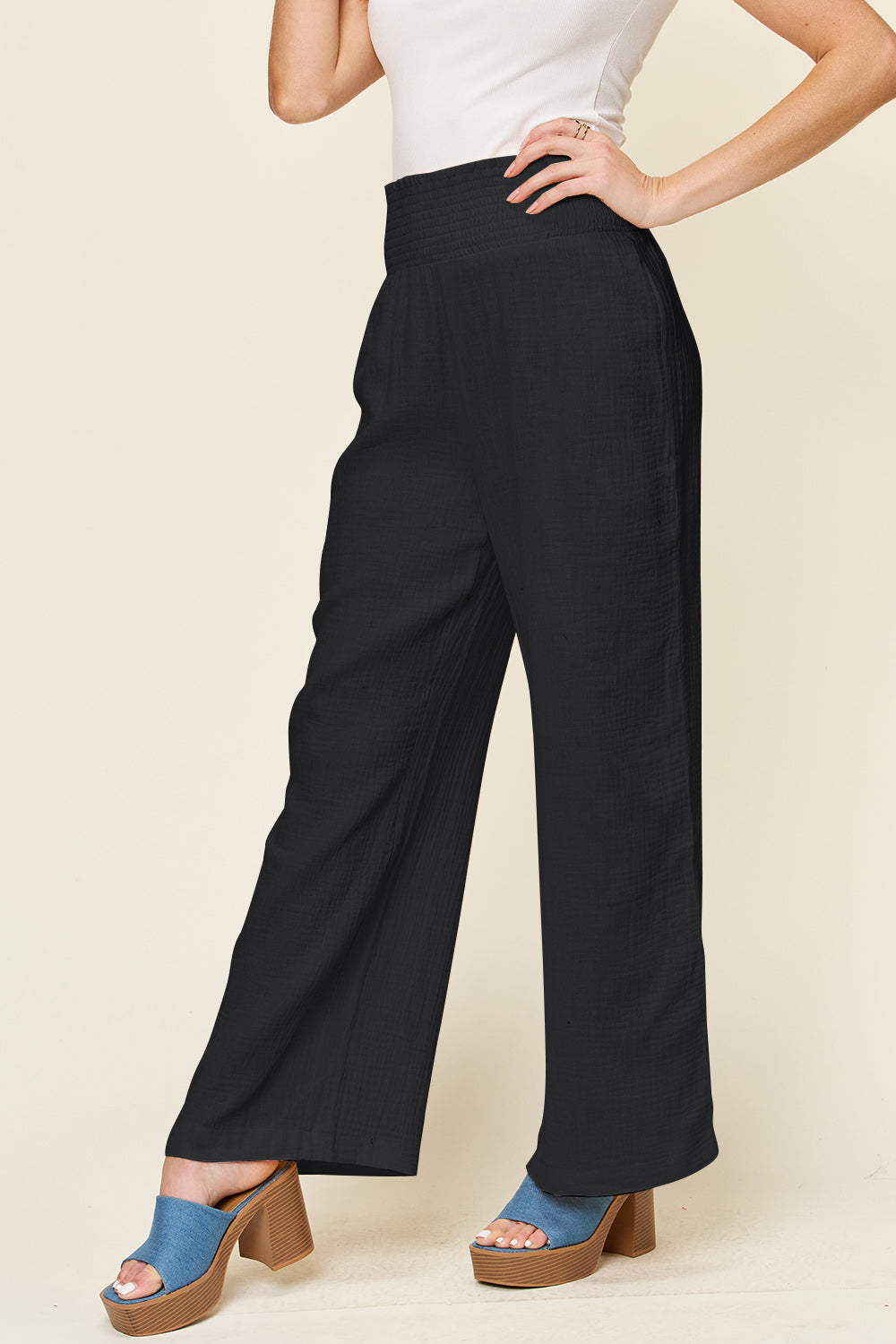 A person wearing Double Take Full Size Texture Smocked Waist Wide Leg Pants in black and a white 100% cotton top is standing with one hand on their hip. They are also wearing blue open-toe sandals.