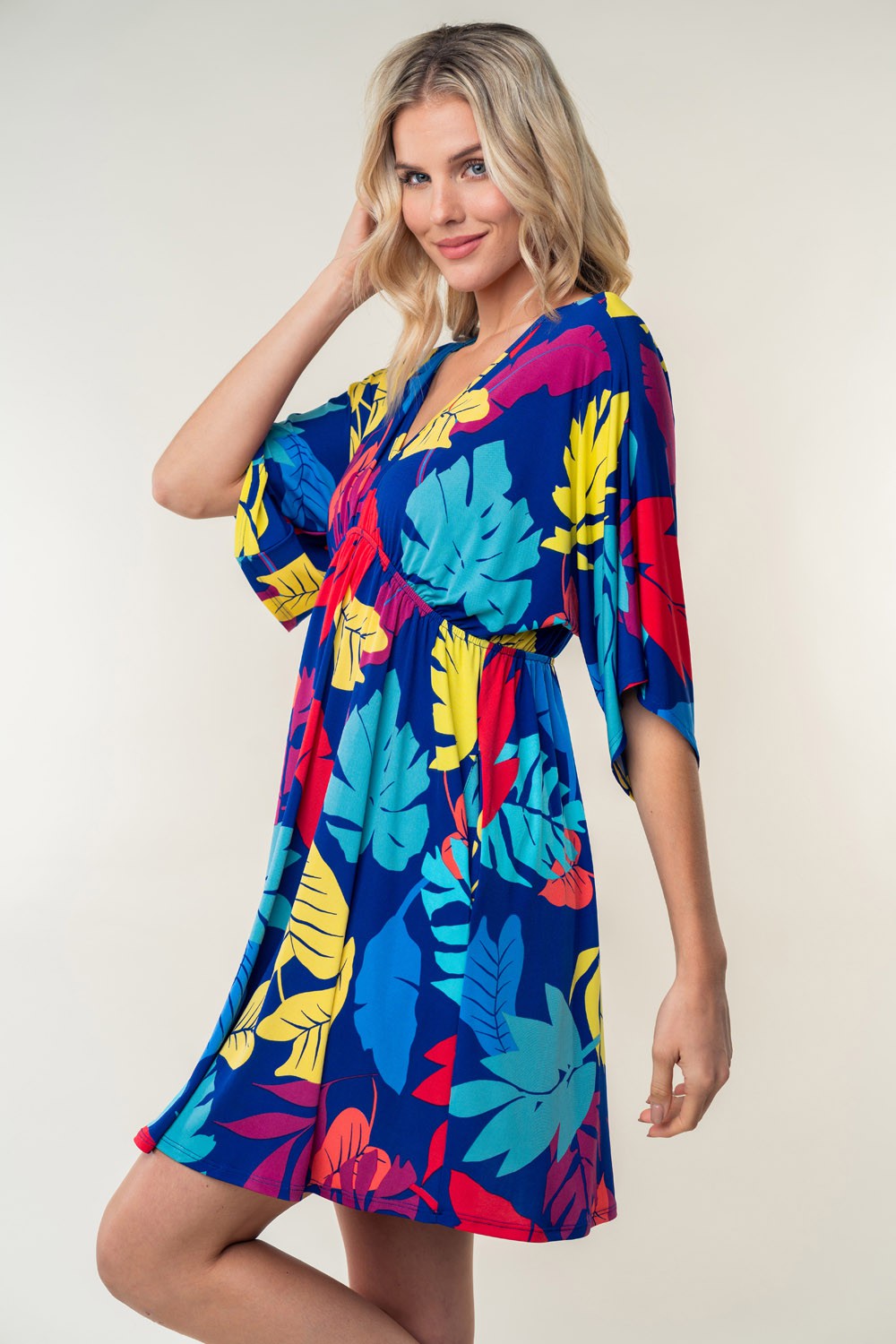 A person wearing the trendy White Birch Full Size Printed V-Neck Mini Dress, adorned with vibrant red, yellow, and blue leaves in a floral pattern, stands against a plain background.