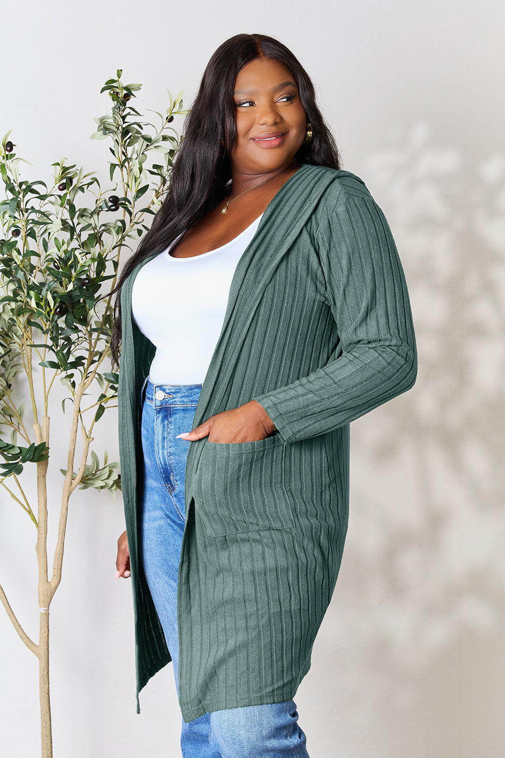 A woman wearing the Basic Bae Full Size Hooded Sweater Cardigan in black, paired with a white top and khaki pants, stands in front of a light background accented by a leafy plant on her left. With one hand tucked into her pocket, she gazes to her right.