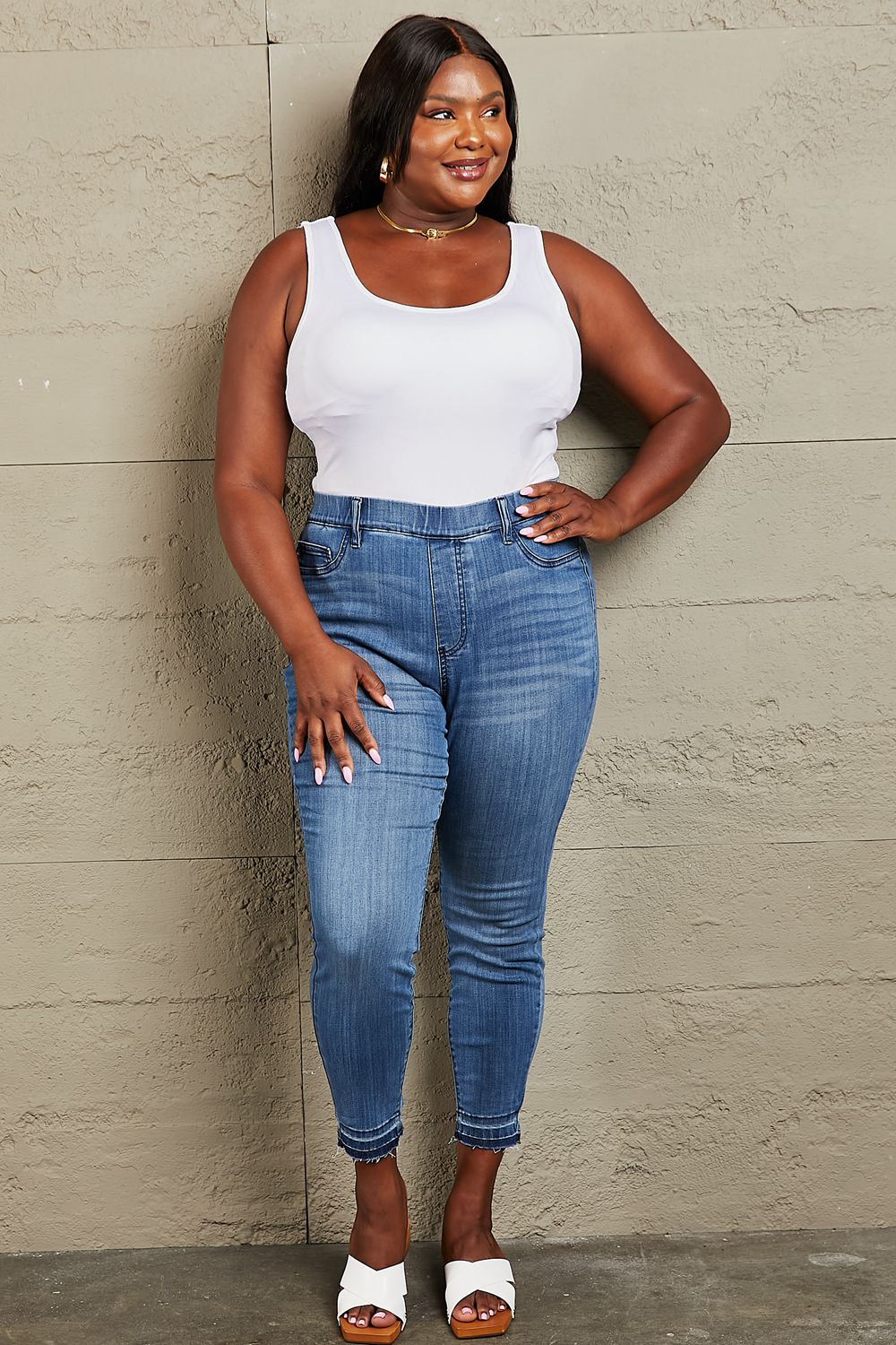 Person wearing Judy Blue Janavie Full Size High Waisted Pull On Skinny Jeans with release hem detailing and white sandals stands against a textured wall.