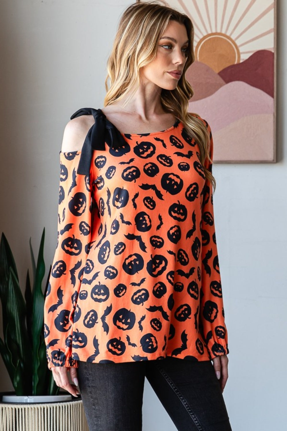 A woman smiles while modeling the Heimish Pumpkin Print One Shoulder T-Shirt, featuring an off-shoulder, long-sleeve design that is perfect for fall season fashion. She stands indoors with a plant and wall art in the background.