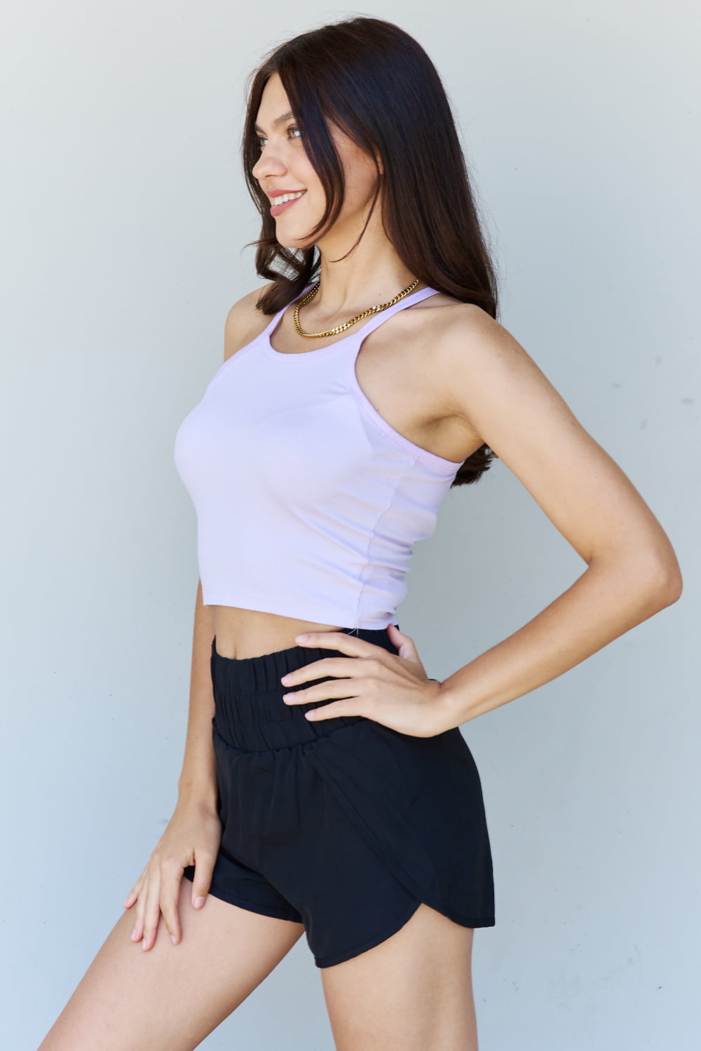 A woman with long brown hair, wearing the Ninexis Everyday Staple Soft Modal Short Strap Ribbed Tank Top in Lavender and black shorts, poses while smiling in front of a plain background.