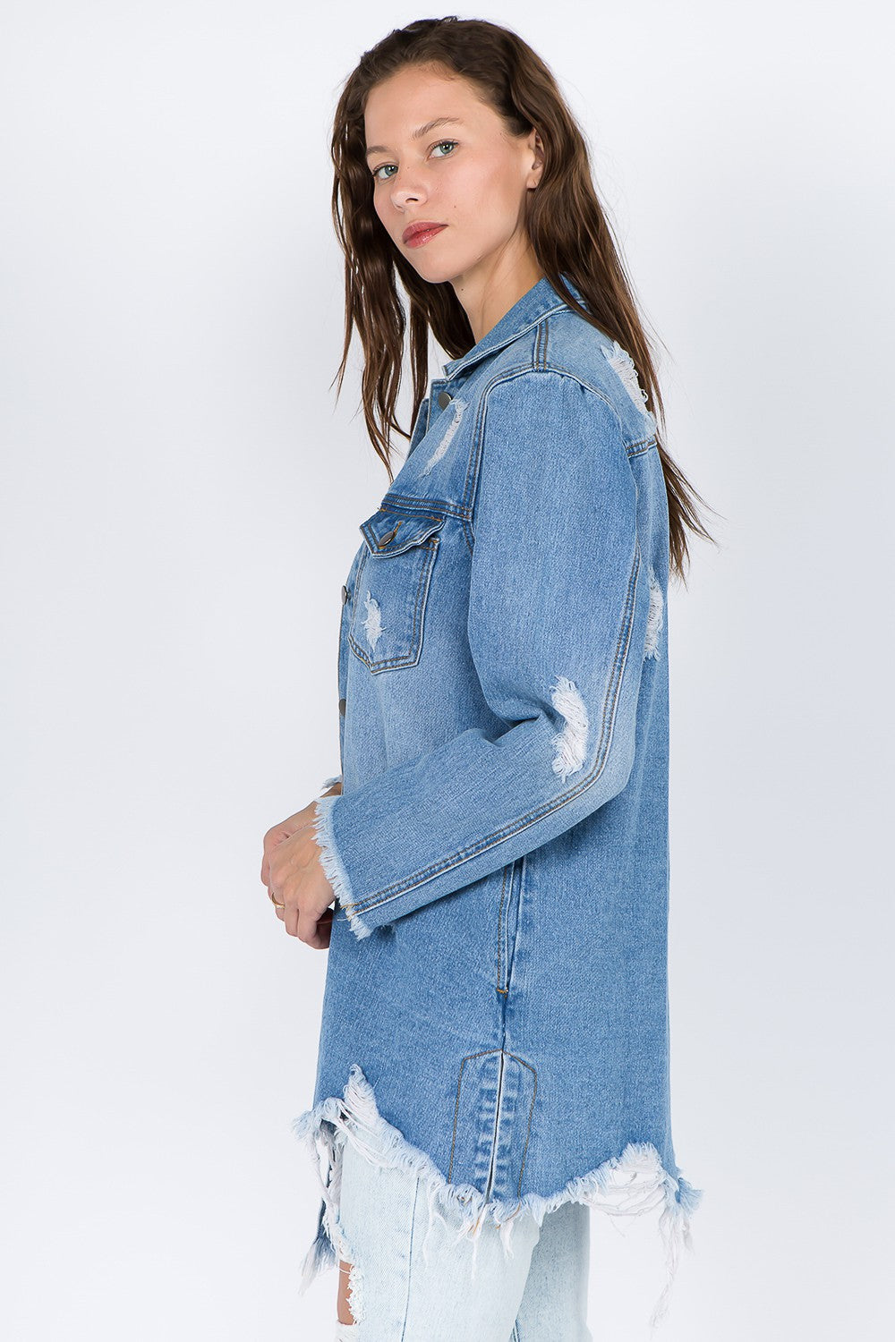 A woman models the American Bazi Distressed Frayed Hem Denim Jacket, featuring frayed edges, a button-up front, and chest pockets that add to its vintage feel. She has long hair and is looking downward with one hand touching her head.