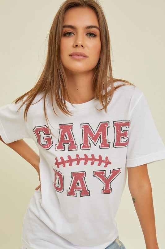 A young woman with long brown hair is wearing a 100% cotton GAME DAY Graphic Tee, featuring a football graphic and "Game Day" text, as she stands against a neutral background.