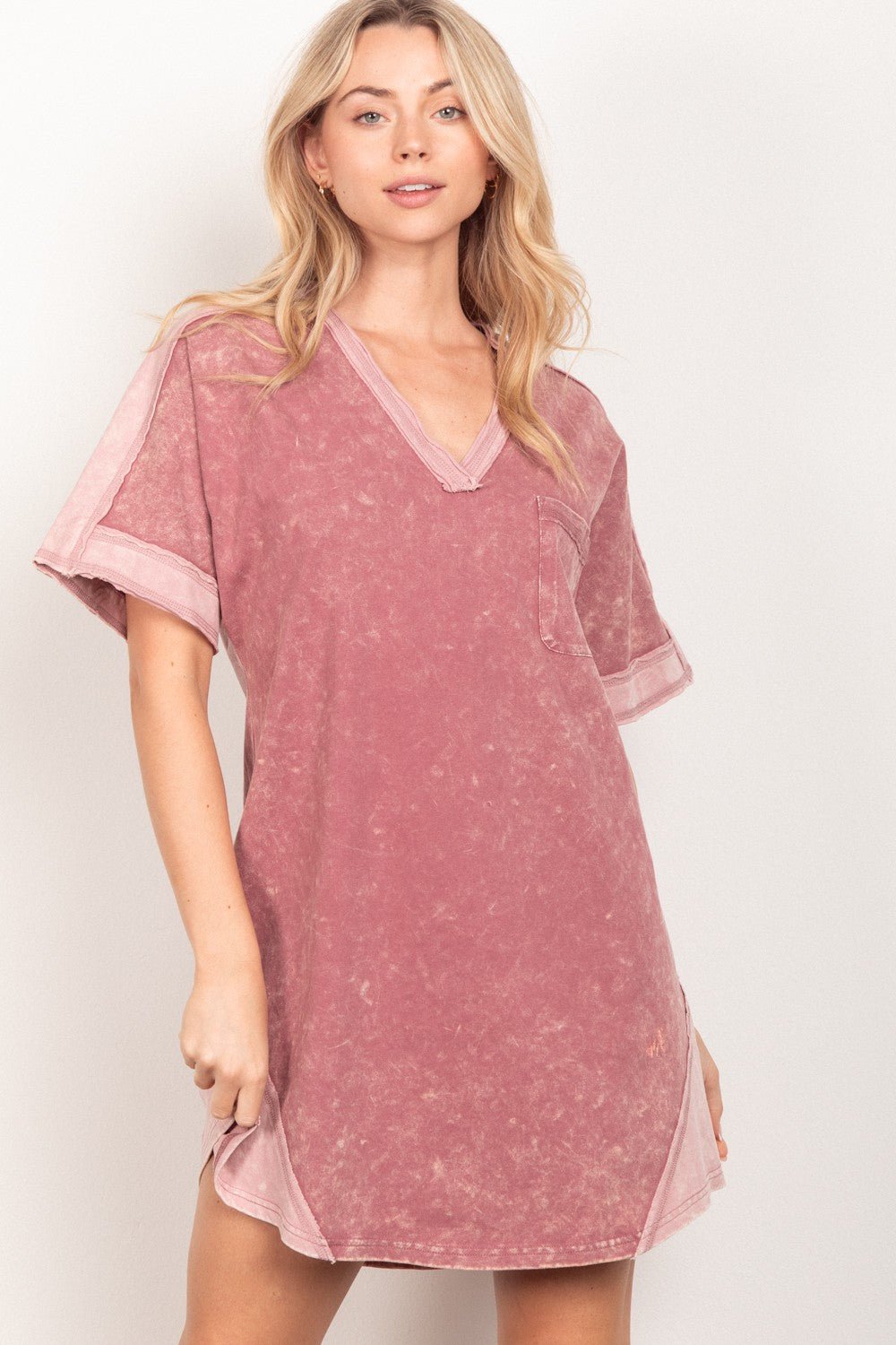 A woman stands against a plain background, wearing the loose-fitting, pink VERY J Short Sleeve V-Neck Tee Dress. Exuding casual style, she gazes at the camera with a slight smile while holding the side of her versatile piece.