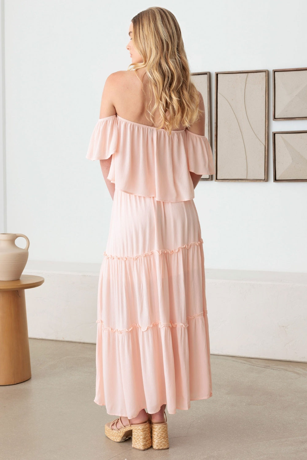 A woman in a romantic Gilli Frill Off-Shoulder Tiered Dress stands holding a beige woven handbag, with two abstract paintings on the wall behind her. She is wearing platform sandals.