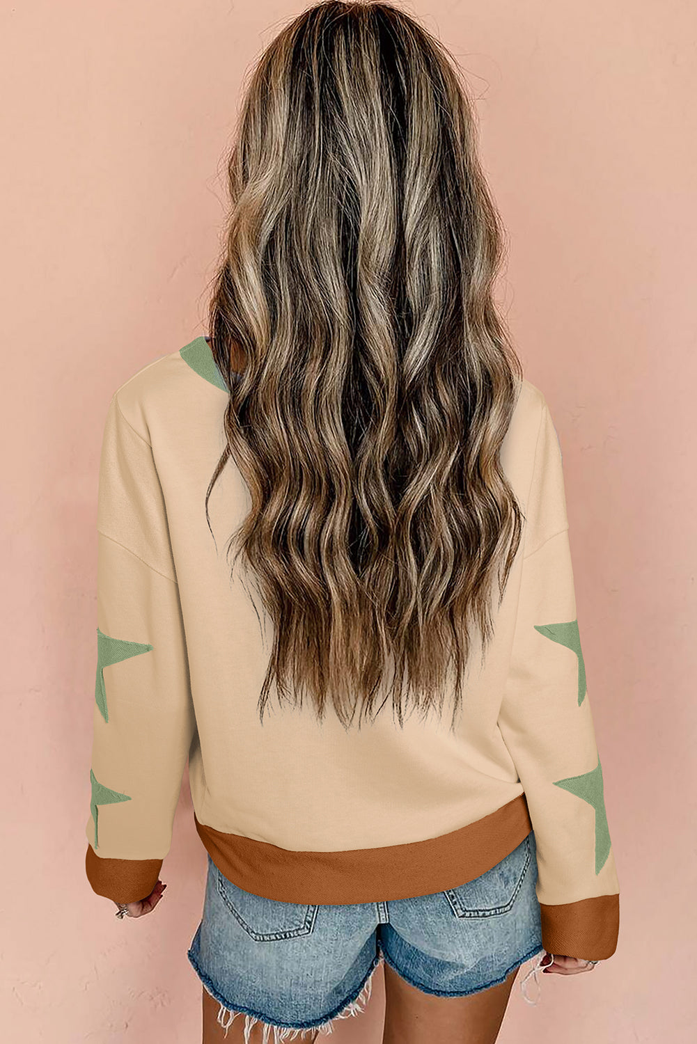 A person with long wavy hair is standing with their back to the camera, wearing a Parchment Star Patchwork Exposed Seam Oversized Sweatshirt and denim shorts. Ideal for a cozy fall/winter wardrobe, this look encapsulates effortless style.