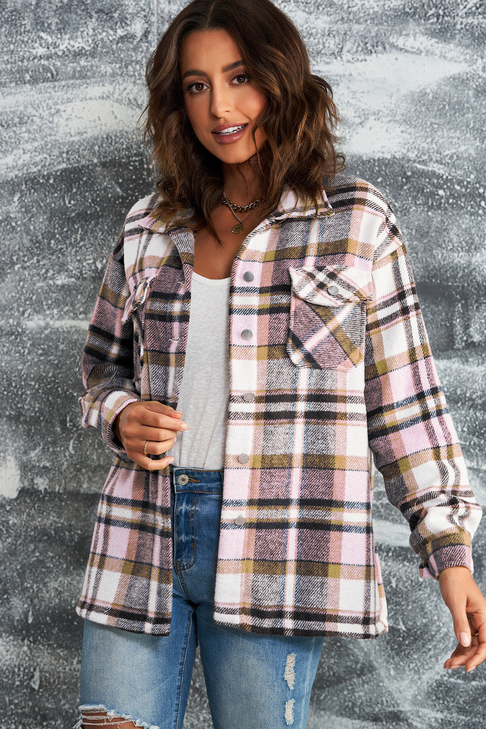 A person wearing a Brown Plaid Button Up Long Sleeve Flannel Shacket layered over a white top with ripped jeans and holding a brown handbag stands against a textured gray wall.