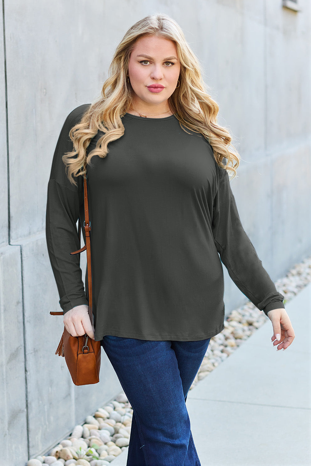 A woman with shoulder-length hair wearing a Basic Bae Full Size Round Neck Dropped Shoulder T-Shirt in green and a pair of jeans, exuding a basic style, stands against a concrete wall. She's holding an imported brown woven backpack on her shoulder while touching her hair with her hand.