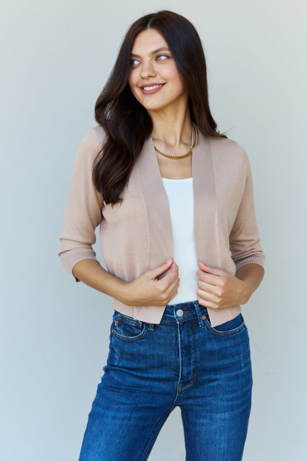 Woman with long dark hair wearing the versatile Doublju My Favorite Full Size 3/4 Sleeve Cropped Cardigan in Khaki over a white top and blue jeans, standing against a plain background.