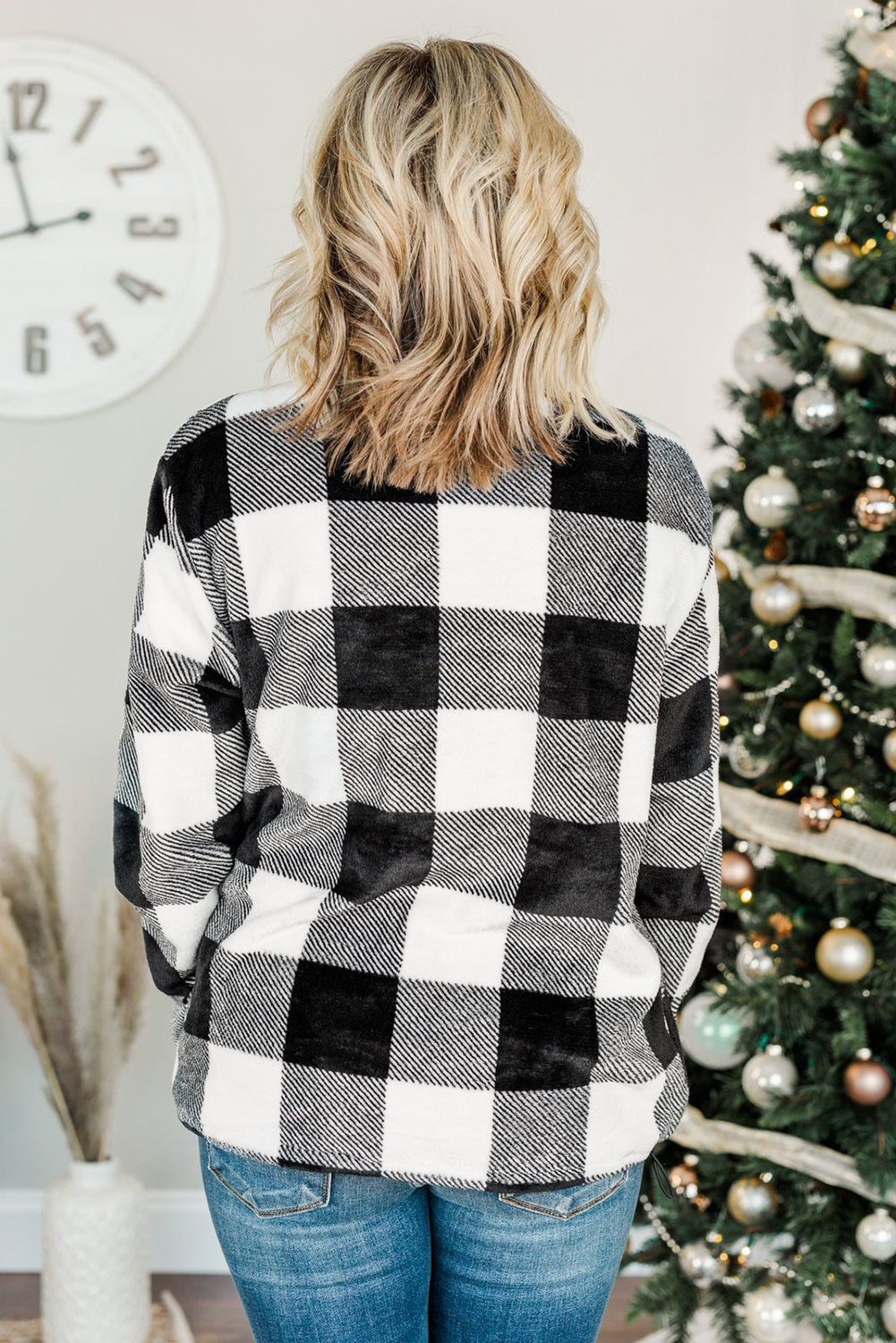 A woman with wavy hair is wearing a cozy Black Plaid Print 1/4 Zip Turn-down Collar Sweatshirt and blue jeans, standing with her back to the camera against a textured gray background.