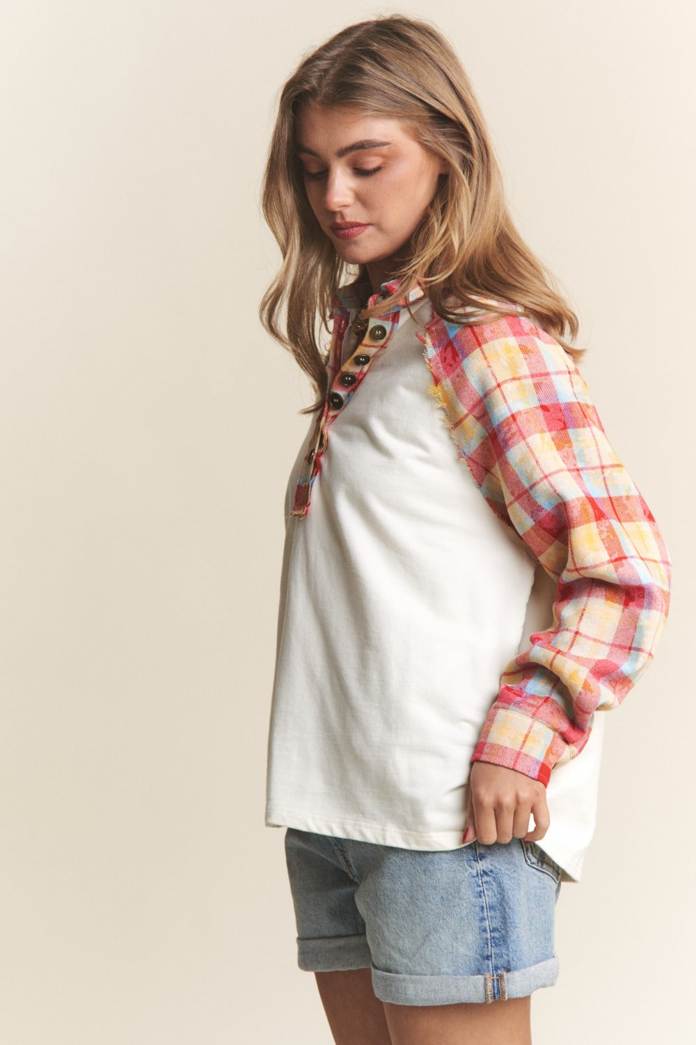 A woman wearing the J.NNA Plaid Contrast Button Down Henley Top—a stylish white pullover shirt with red and yellow plaid sleeves—and denim shorts, stands against a plain background.