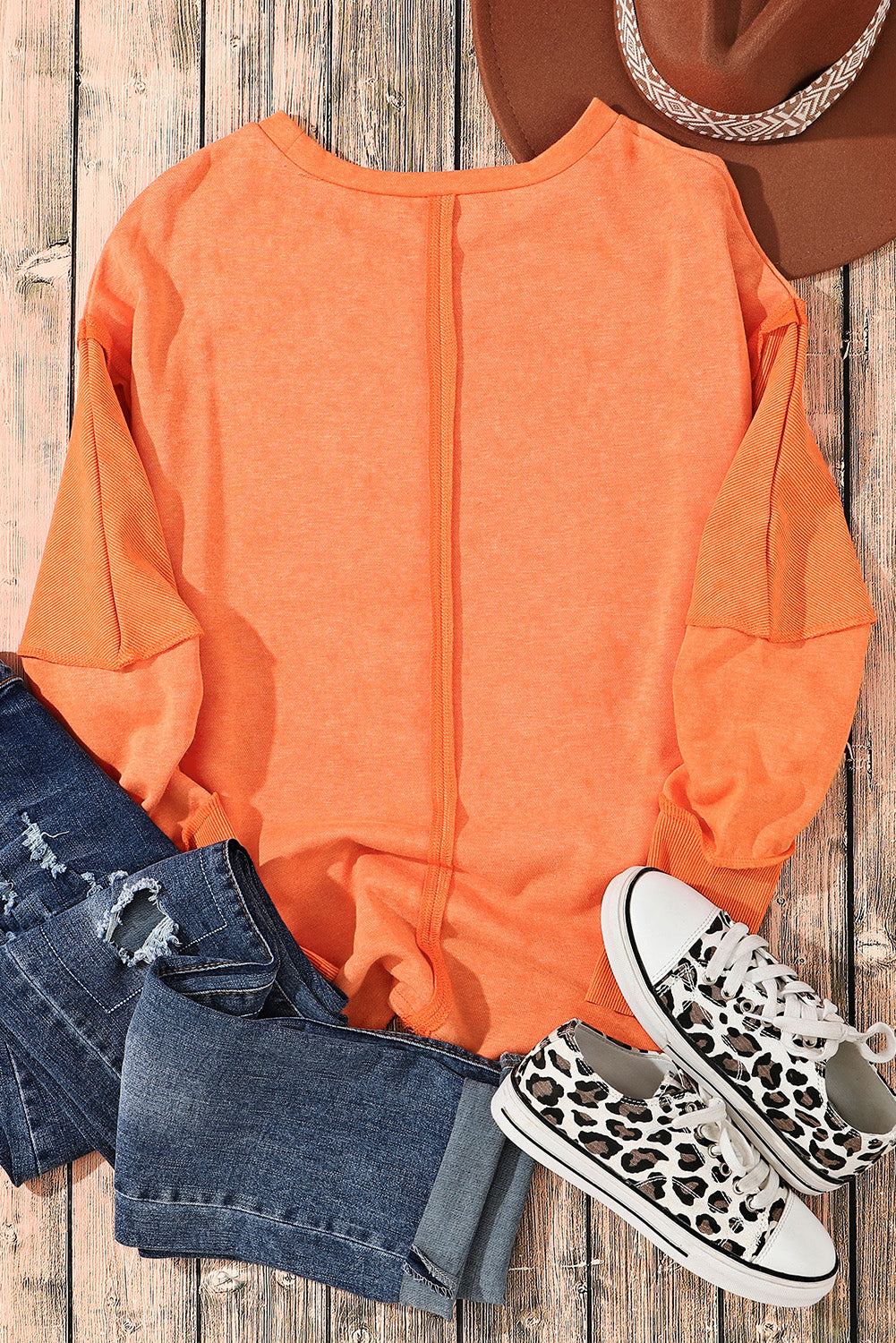 A person with long blonde hair is standing with their back to the camera, wearing an Orange Exposed Seamed High Low Raw Edge Sweatshirt and denim shorts, in front of a brick wall.