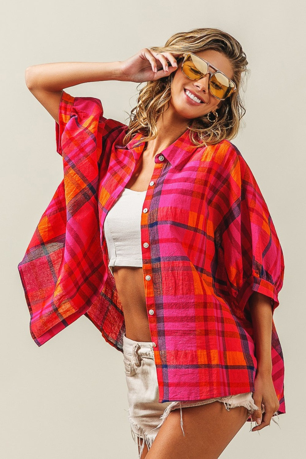 A woman outfitted in a BiBi Button Up Dolman Sleeve Plaid Shirt, paired with a white crop top, distressed denim shorts, and yellow sunglasses.