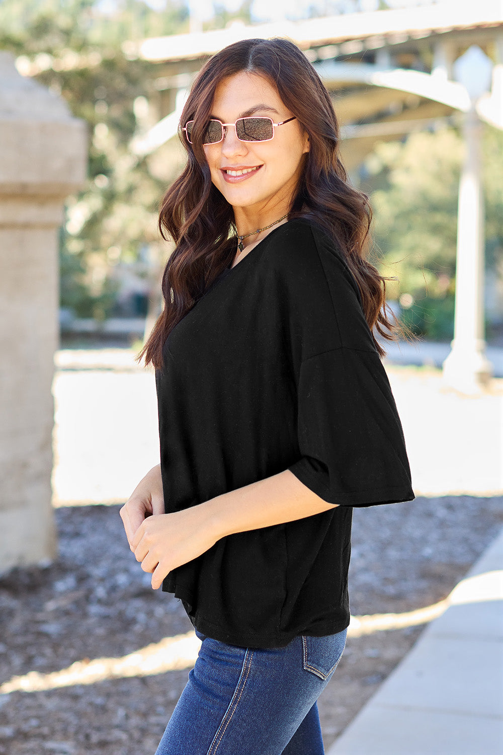 A person with long dark hair and a bright smile stands outdoors, showcasing their basic style in a loose, slightly stretchy *Basic Bae Full Size Round Neck Drop Shoulder T-Shirt* in pink and blue jeans, with one hand on their hip.