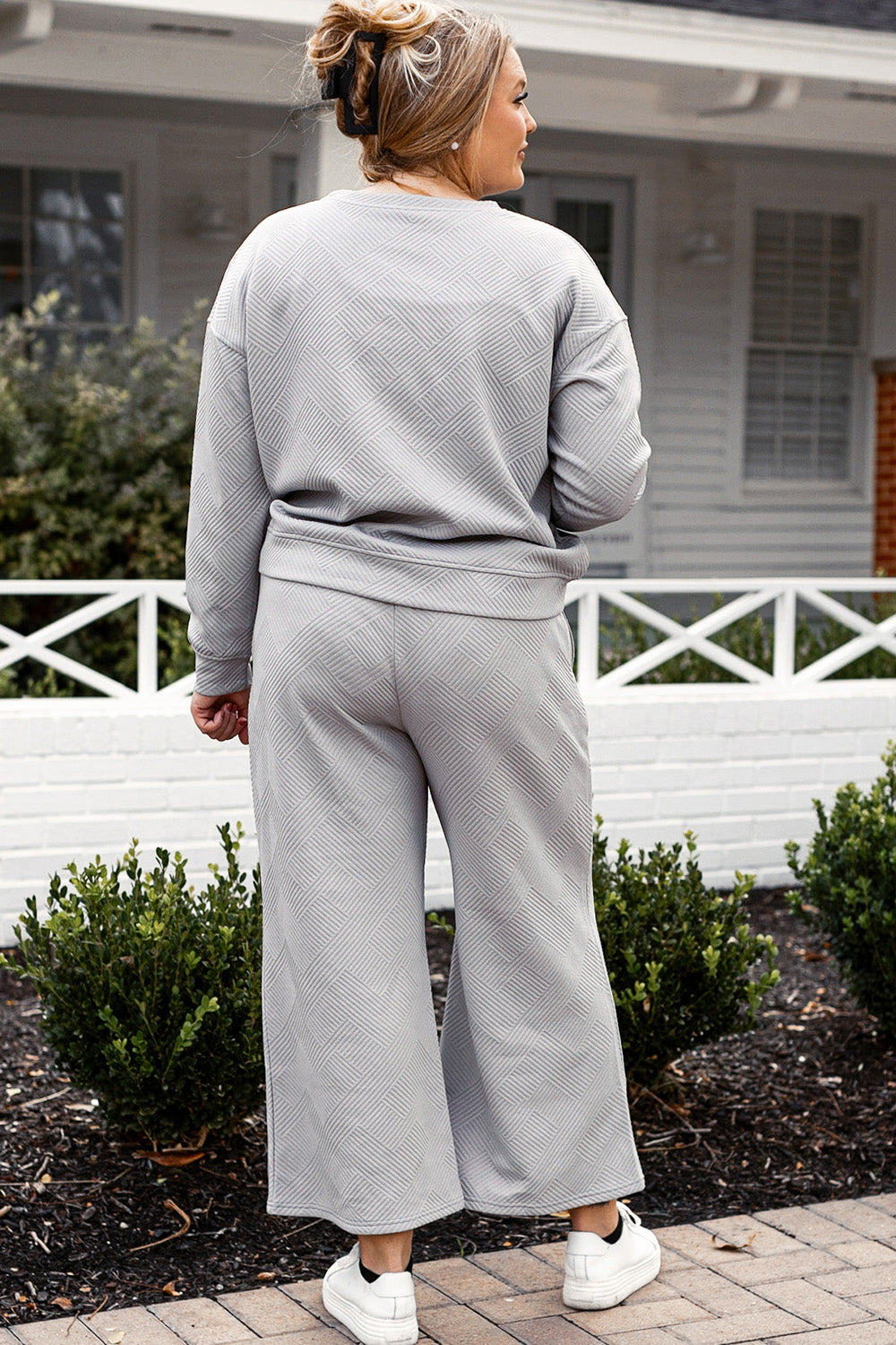 A woman with blonde hair, dressed in the Double Take Full Size Textured Long Sleeve Top and Drawstring Pants Set featuring a stretchy black long-sleeve top and matching wide-leg trousers made of rayon spandex, stands barefoot in a room with white walls and a wooden floor.