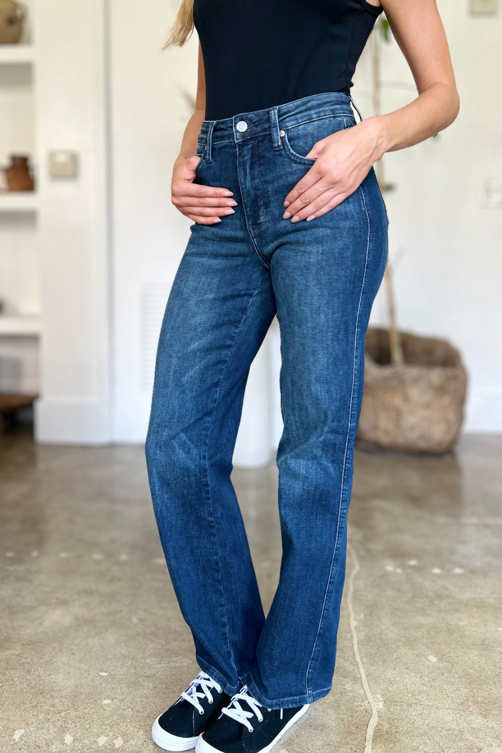 A person wearing a black top and Judy Blue Full Size Tummy Control Straight Jeans stands indoors with one hand on their hip. Their shoes are black with white soles. A plant and shelves are in the background.