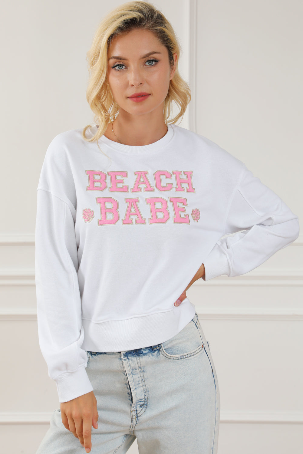 A person with long blonde hair is standing with their back to the camera, wearing a White BEACH BABE Slogan Graphic Casual Sweatshirt and light blue jeans.