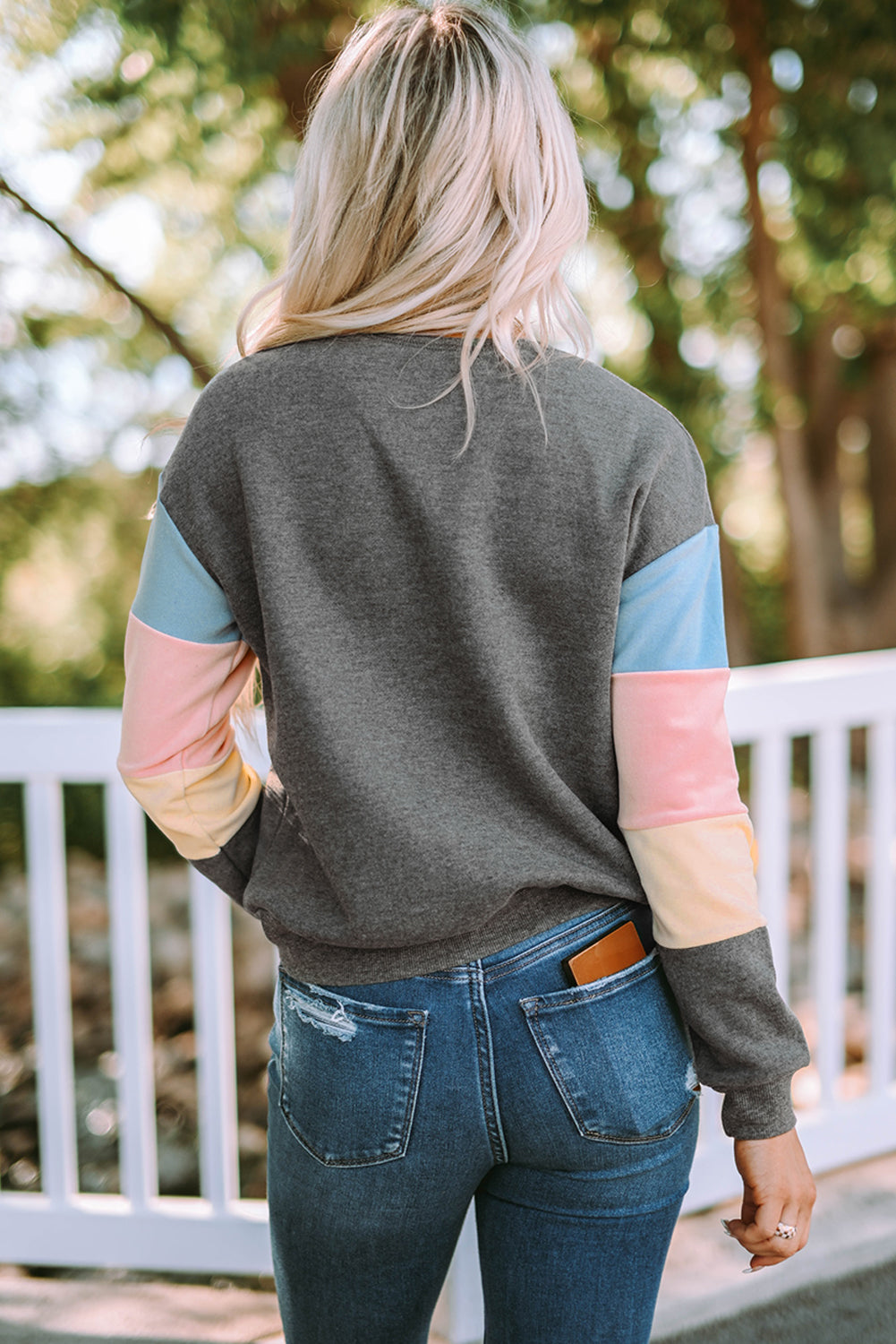 A person with long blonde hair wearing a stylish Gray Colorblock Long Sleeve Pullover Sweatshirt and blue jeans stands outdoors near a white fence.