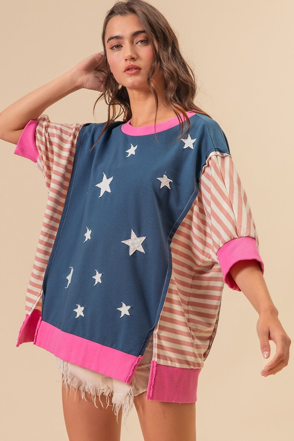 A person wearing a BiBi US Flag Theme Color Block Star Patch T-Shirt, characterized by its oversized fit, blue base with white star patterns, and pink-striped sleeves, stands against a plain background. They have their hand touching their hair.