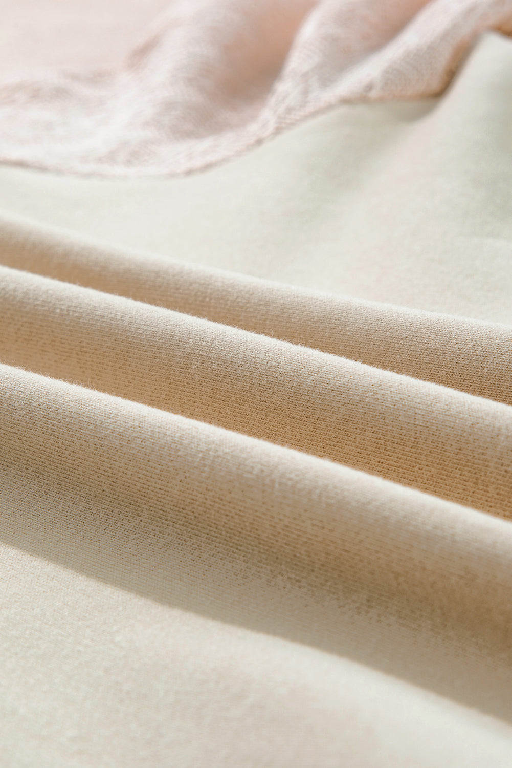 A blonde-haired individual wearing a Parchment Eyelet Knit Patchwork Raglan Sleeve Pullover Top stands indoors, smiling and looking to the side. Flowers and a ladder can be seen in the background.