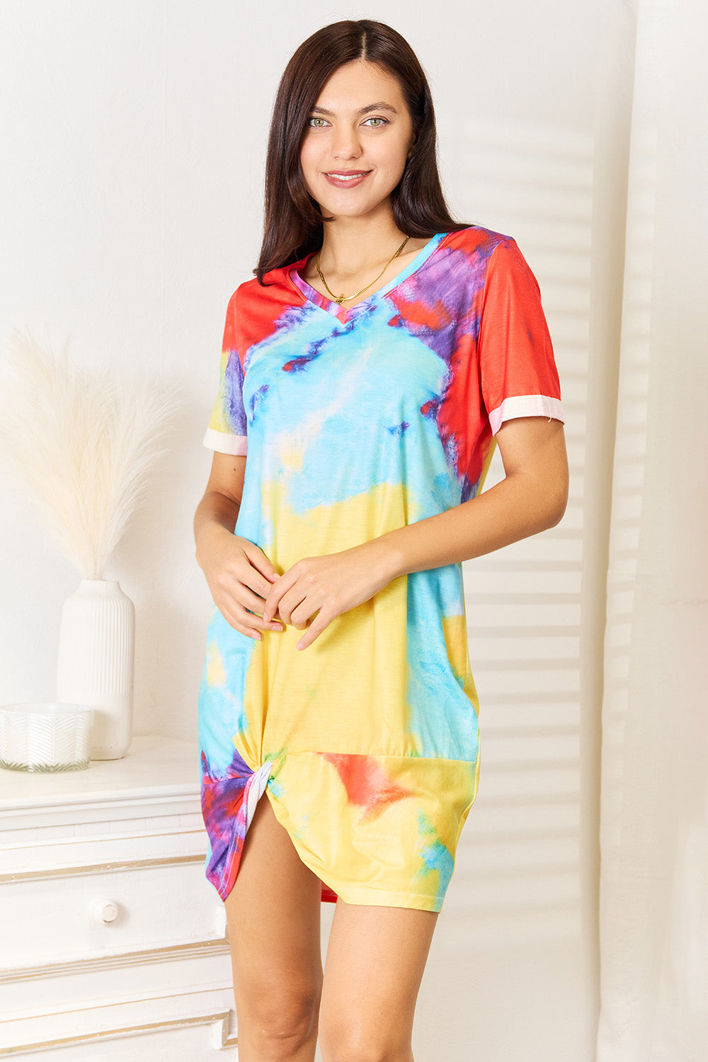 A person wearing the Shiny Tie-Dye V-Neck Twisted Dress stands indoors near a white dresser, smiling with hands clasped in front.