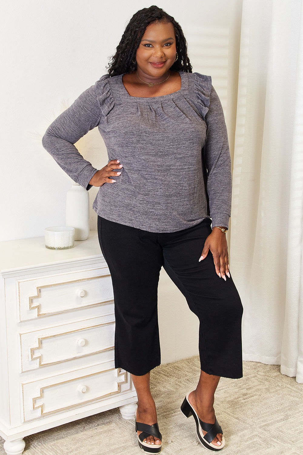 A woman wearing a Double Take Square Neck Ruffle Shoulder Long Sleeve T-Shirt in pink and blue jeans stands with one hand in her pocket, smiling. She has long curly hair and wears a necklace and bracelet. She stands in a room with white decor.