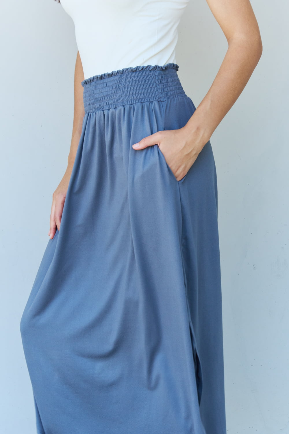 A person wearing a white sleeveless top and the Doublju Comfort Princess Full Size High Waist Scoop Hem Maxi Skirt in Charcoal stands against a plain wall. They are also wearing black sandals.