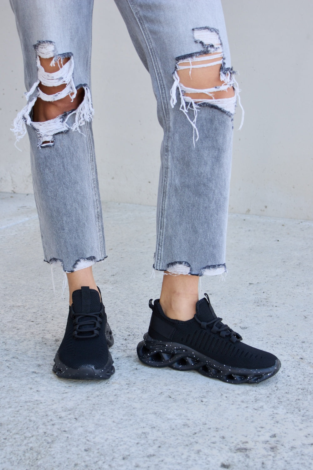 A person wearing ripped gray jeans and Forever Link Mesh Breathable Lace-Up Athletic Shoes is standing on a concrete surface.