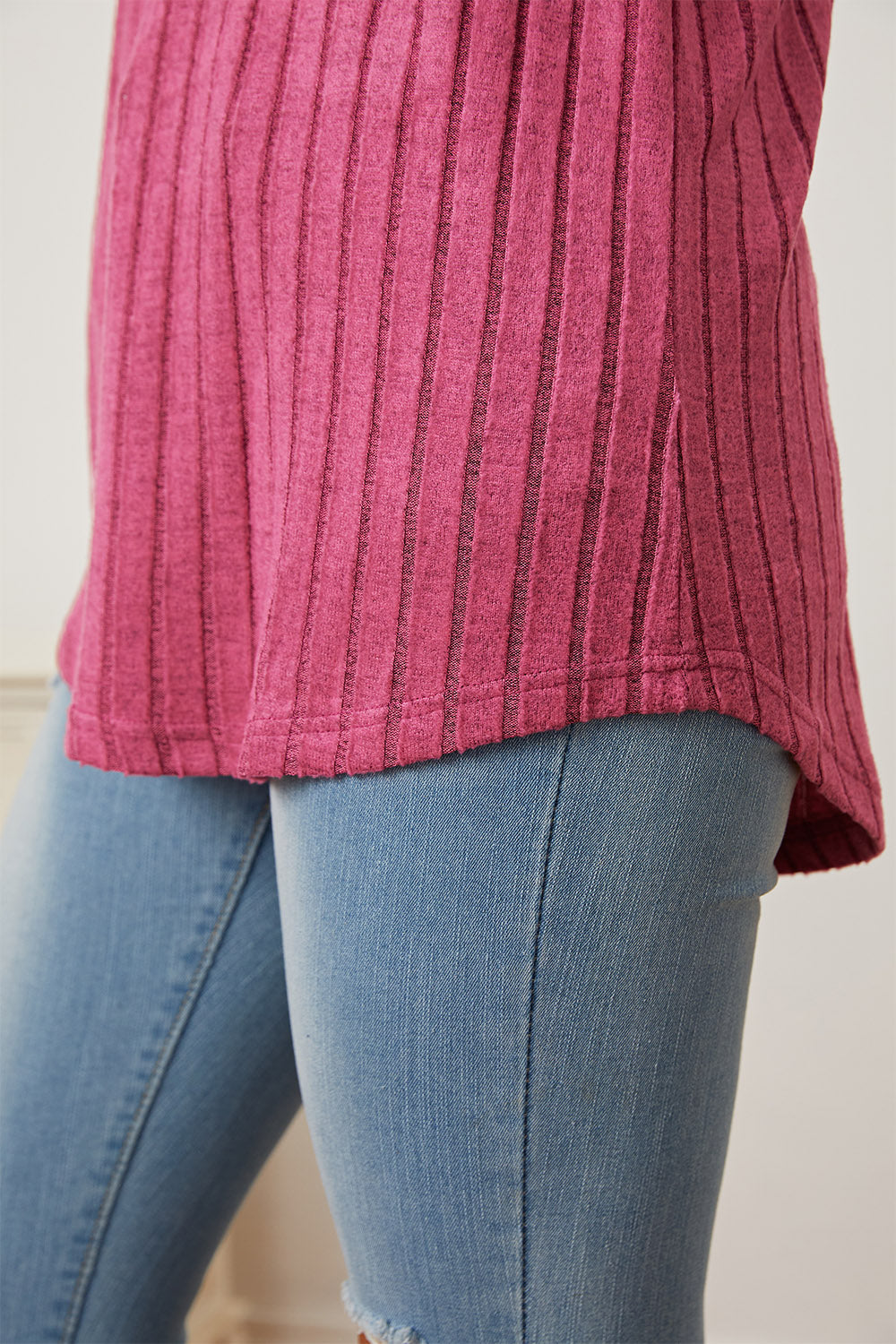 A person wearing the Basic Bae Full Size Ribbed Thumbhole Sleeve T-Shirt in blue and light-washed jeans stands smiling, with a decorative wall hanging in the background.