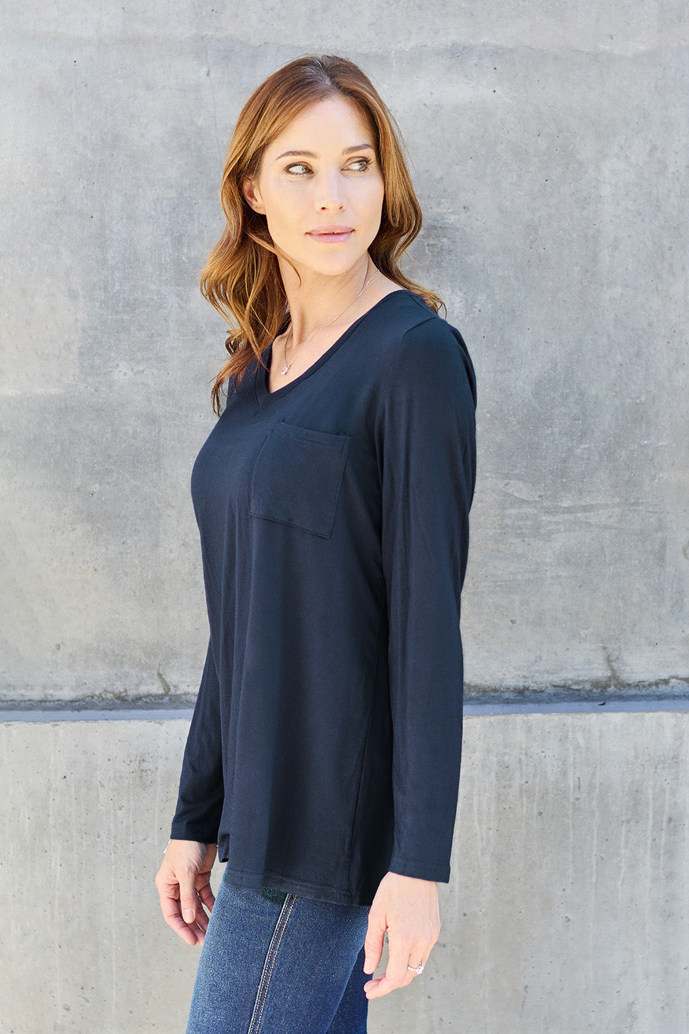 A woman with light brown hair stands against a concrete wall, wearing the Basic Bae Full Size V-Neck Long Sleeve Top in olive green and blue jeans, with a white backpack on her shoulder. Her outfit exudes basic style and is made from polyester, ensuring it’s both comfortable and suitable for machine wash cold. She is looking at the camera.