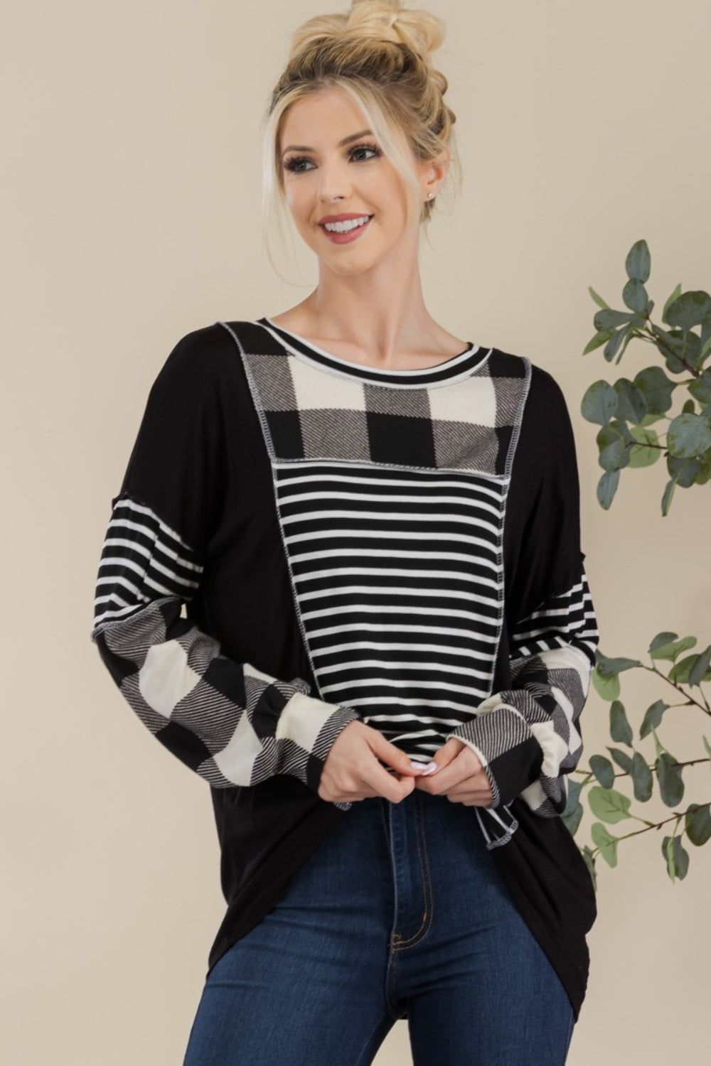 A person wearing a Celeste Full Size Color Block Exposed Seam T-Shirt, featuring black and white plaid with reverse stitching and long sleeves, poses while smiling in front of a neutral background with a plant.