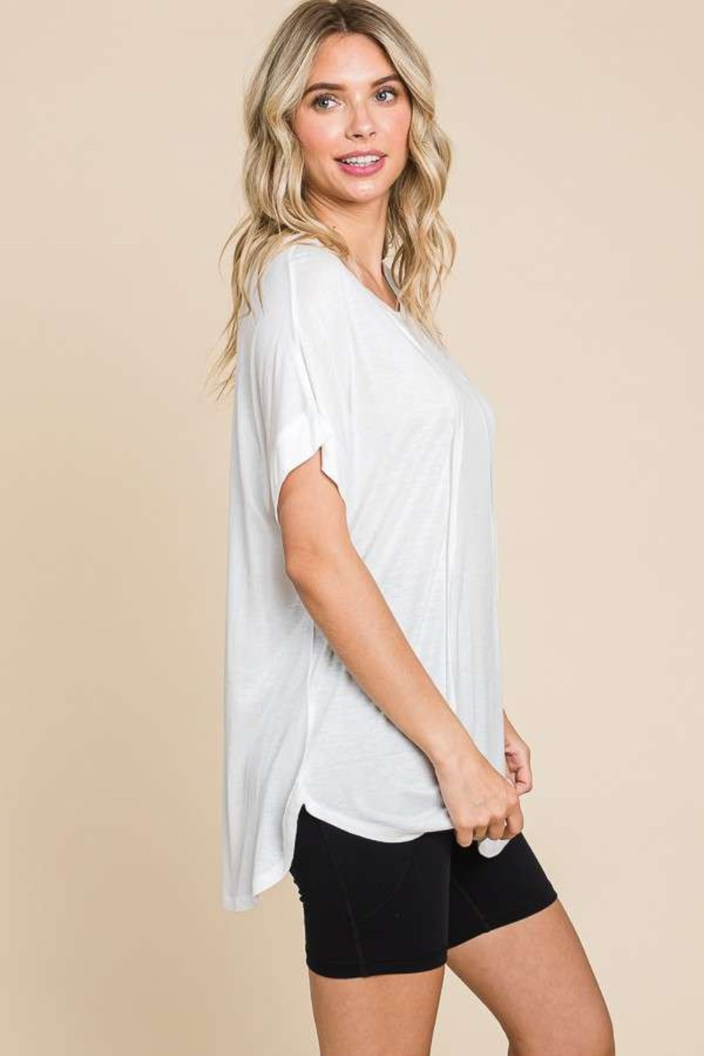 Woman in a white Culture Code Round Neck Short Sleeve T-Shirt and black shorts, posing against a beige background with one hand on her hip. This versatile staple makes everyday wear effortlessly chic.