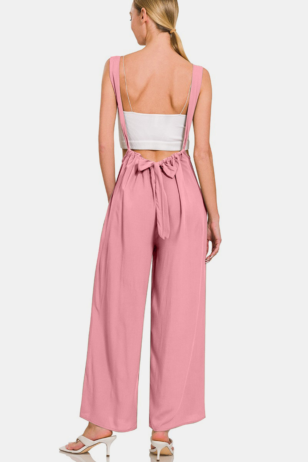 A woman is smiling in her fashion-forward style, donning the Zenana Pocketed Wide Strap Wide Leg Overalls in pink along with a white crop top. She has her hands in the overall's pockets and complements the look with white high-heeled shoes.