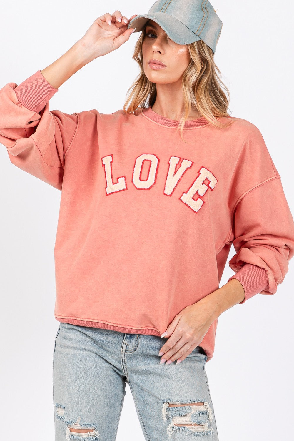 A person with long hair wears a cap, an oversized pink SAGE + FIG LOVE Path Applique Drop Shoulder Sweatshirt, and ripped jeans, standing against a plain background.