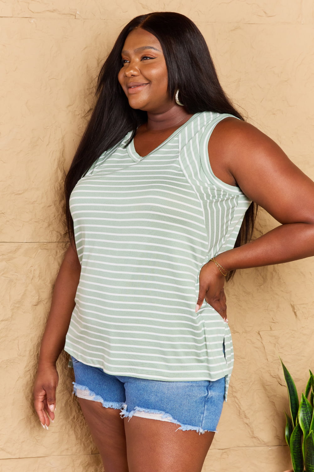 A person wearing the Doublju Full Size Striped Sleeveless V-Neck Top paired with denim shorts stands against a textured beige wall, smiling and posing with one hand on their hip.