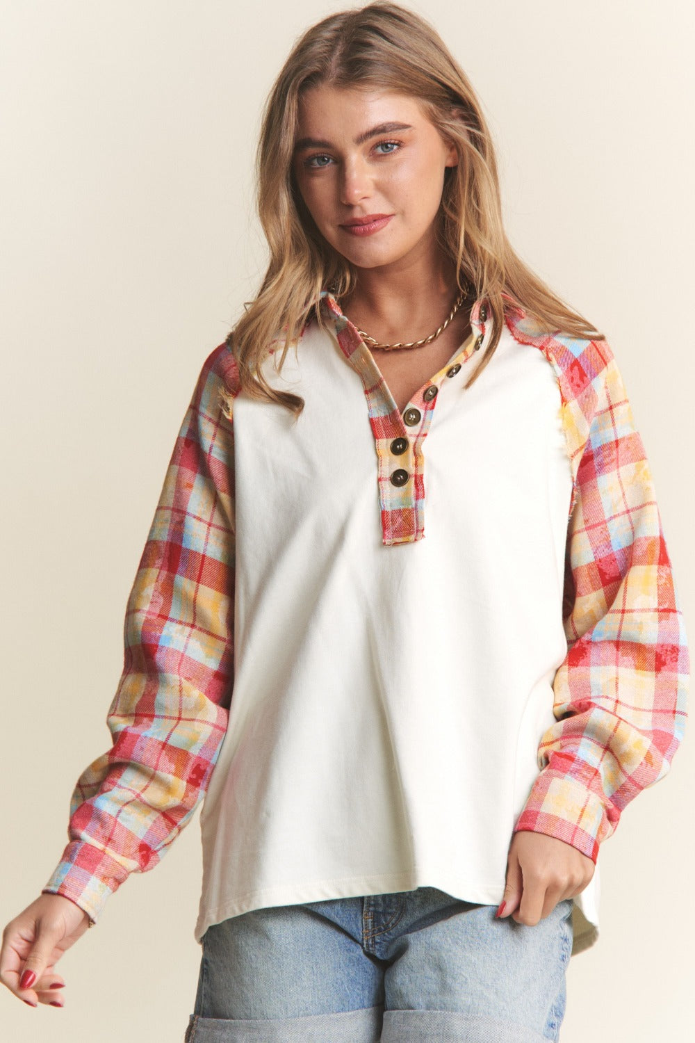 A woman wearing the J.NNA Plaid Contrast Button Down Henley Top—a stylish white pullover shirt with red and yellow plaid sleeves—and denim shorts, stands against a plain background.