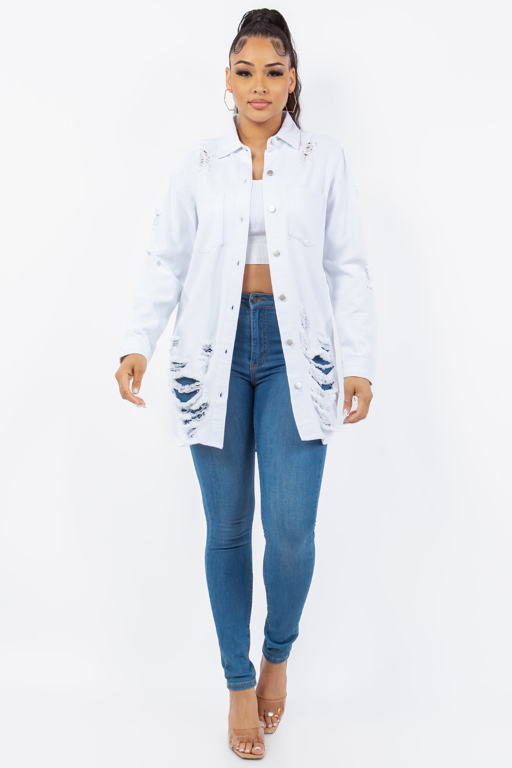 A woman wearing an American Bazi Distressed Button Up Long Sleeve Denim Jacket over a white crop top and blue jeans stands facing the camera against a plain background.