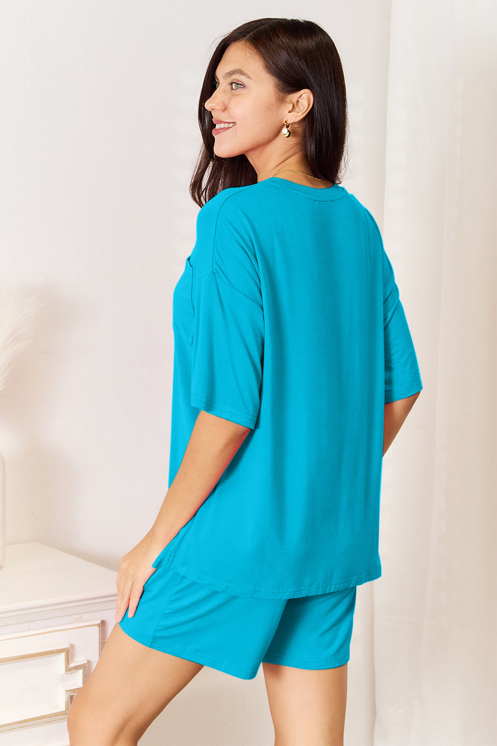 A woman is standing in a brightly lit room, wearing the Basic Bae Full Size Soft Rayon Half Sleeve Top and Shorts Set in a relaxed, stretchy turquoise fabric. She has one hand placed in her pocket and is smiling while looking slightly to the side.