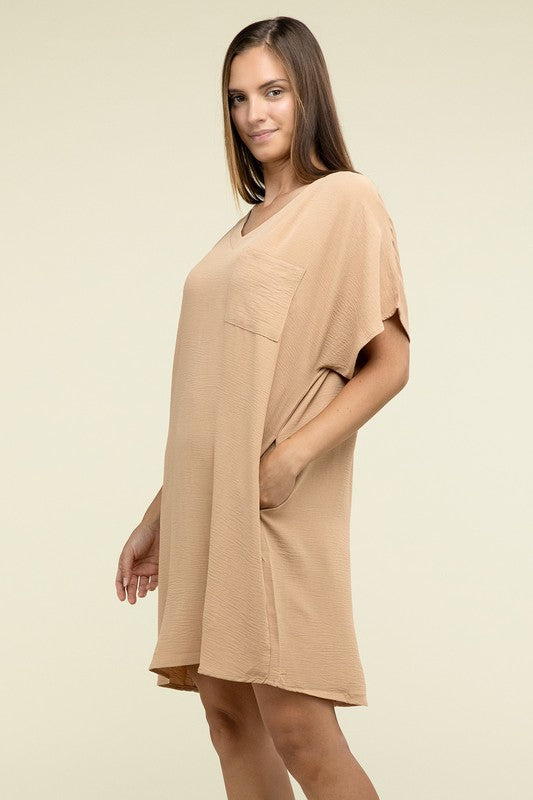 A woman wearing a loose light brown Woven Airflow V Neck T-Shirt Dress with Pockets stands against a plain background. Made from breathable fabric, this summer dress includes pockets where her hands rest as she smiles slightly.