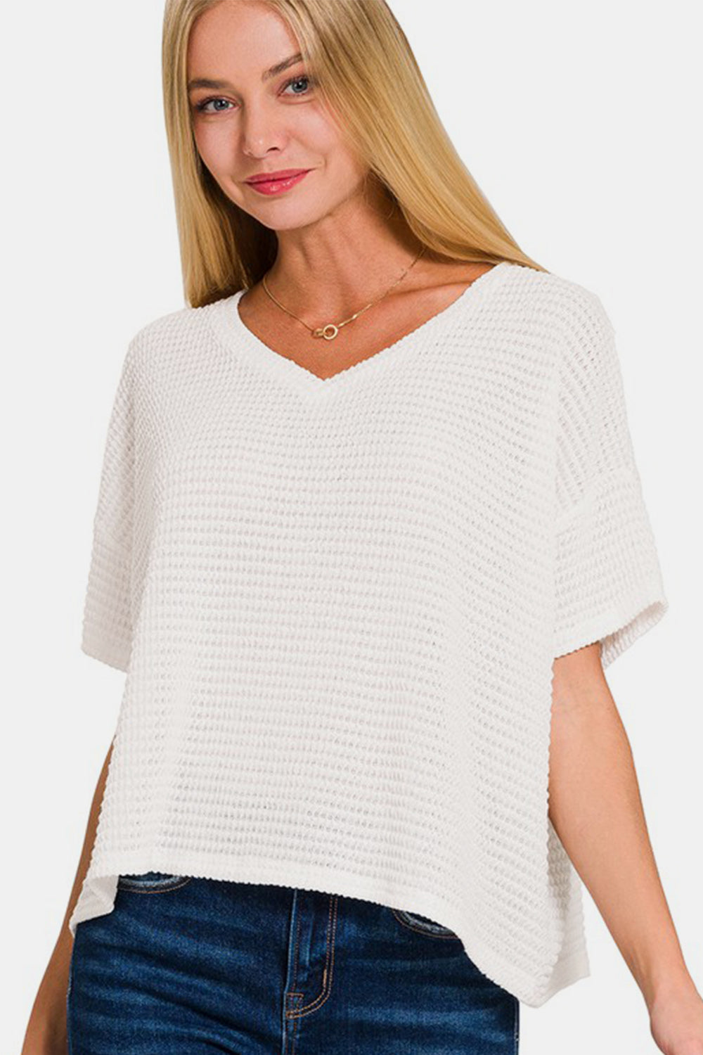 A woman wearing a Zenana Drop Shoulder Short Sleeve Jacquard Knit Top in white stands with one hand on her hip and smiles. She has long, light brown hair and is paired with dark blue jeans.