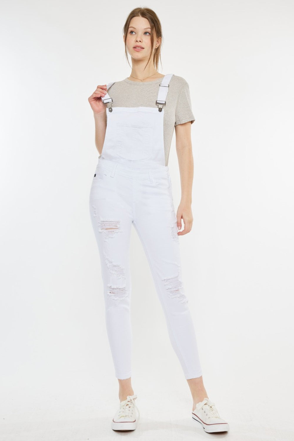 A fashion-forward woman stands against a plain background, wearing a grey t-shirt, Kancan Distressed Skinny Denim Overalls in white, and white sneakers with red detailing.