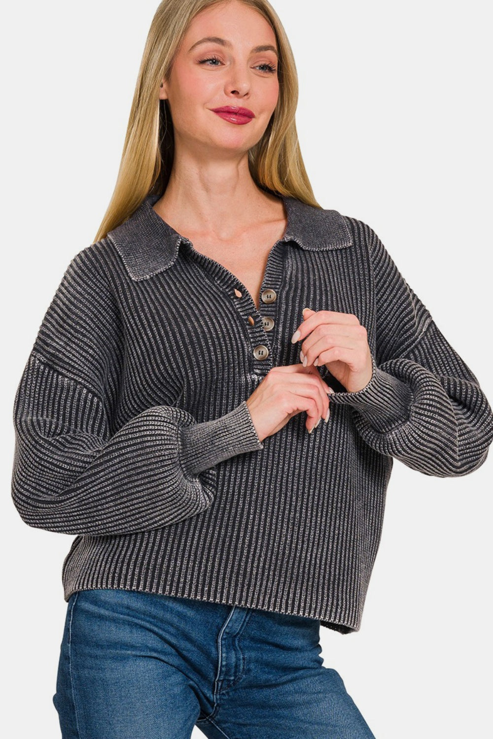 A woman with long blonde hair modeling the Zenana Washed Half Button Long Sleeve Sweater, made of soft washed fabric in grey, paired with blue jeans against a plain backdrop.