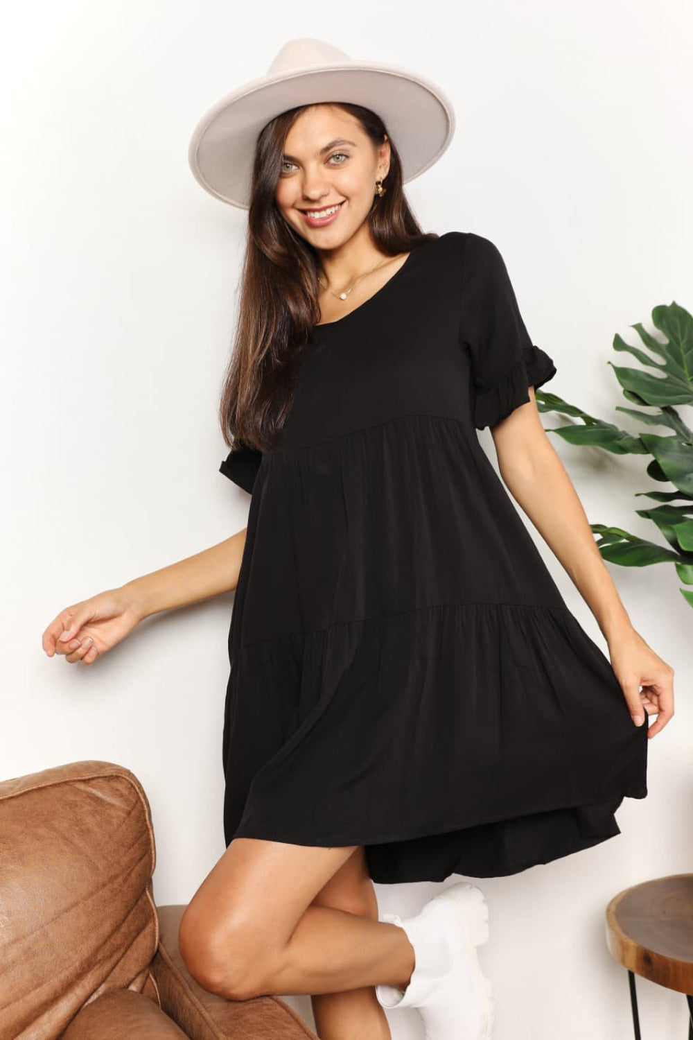 A woman stands indoors wearing the Mandy V-Neck Flounce Sleeve Tiered Dress in navy blue. She has one hand on her hip and is smiling.