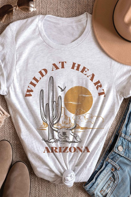 Woman wearing the WILD AT HEART ARIZONA Graphic Tee in white, which features the text "Wild at Heart Arizona" along with an illustration of a cactus, a sun, and a lounging skeleton. This tee is made from 100% cotton and offers unisex sizing for ultimate comfort and style.