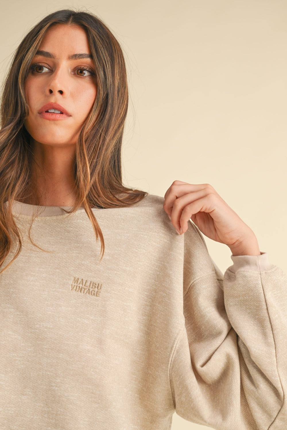 A woman with long brown hair wears the Mable Round Neck Letter Embroidery Crop Sweatshirt in beige, paired with blue jeans, standing confidently with one hand in her pocket against a plain background, perfectly capturing effortless street style.