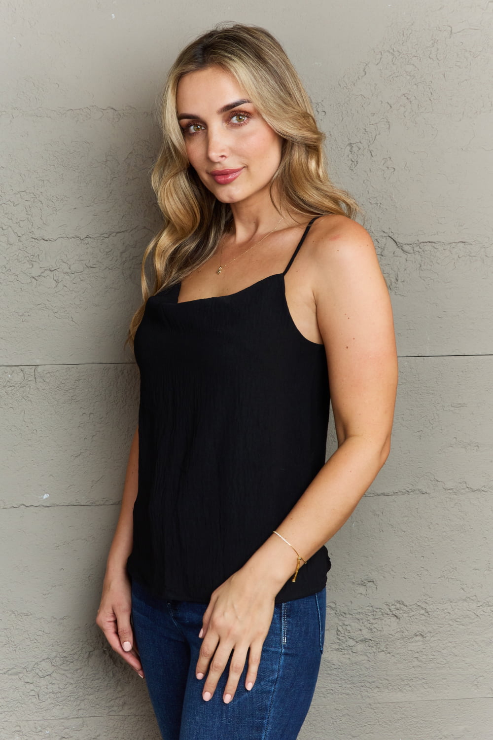 A person with long wavy hair, wearing the Ninexis For The Weekend Loose Fit Cami in black and jeans, stands smiling against a textured gray wall.