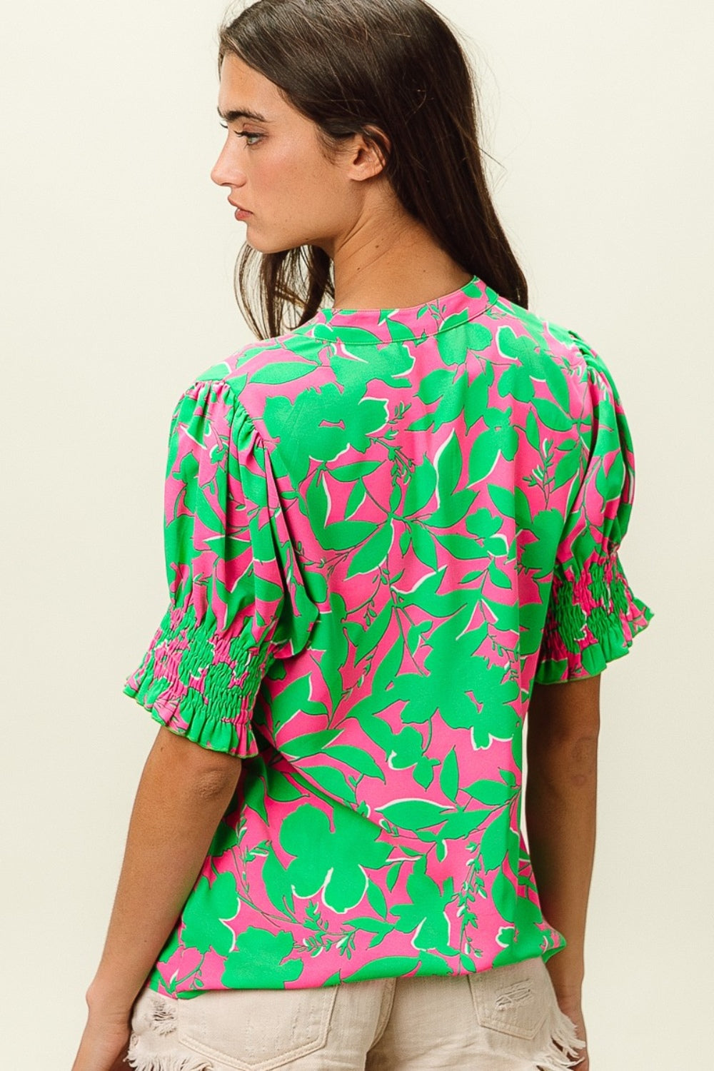 A woman wearing the BiBi Smocked Cuff Floral Contrast Top, featuring a chic design with bright pink and green floral patterns and ruffled sleeves, poses with one hand near her chin.