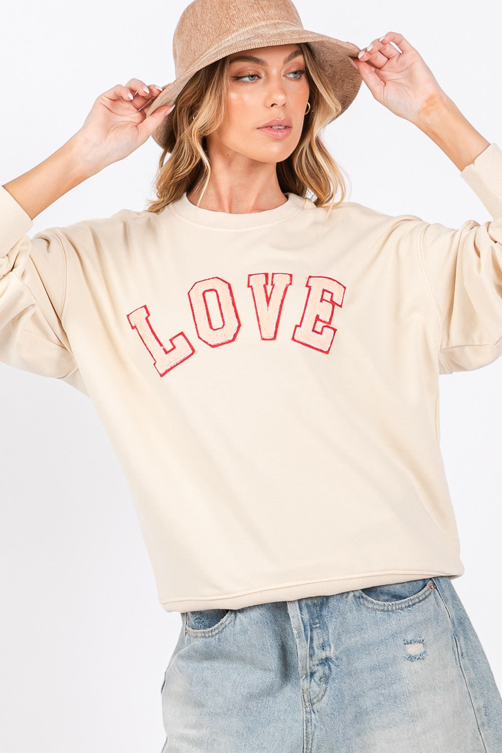 A woman dressed in a SAGE + FIG LOVE Path Applique Drop Shoulder Sweatshirt and a wide-brimmed hat is touching the brim of her hat with both hands, looking to the side and flaunting her cozy and stylish sweatshirt.